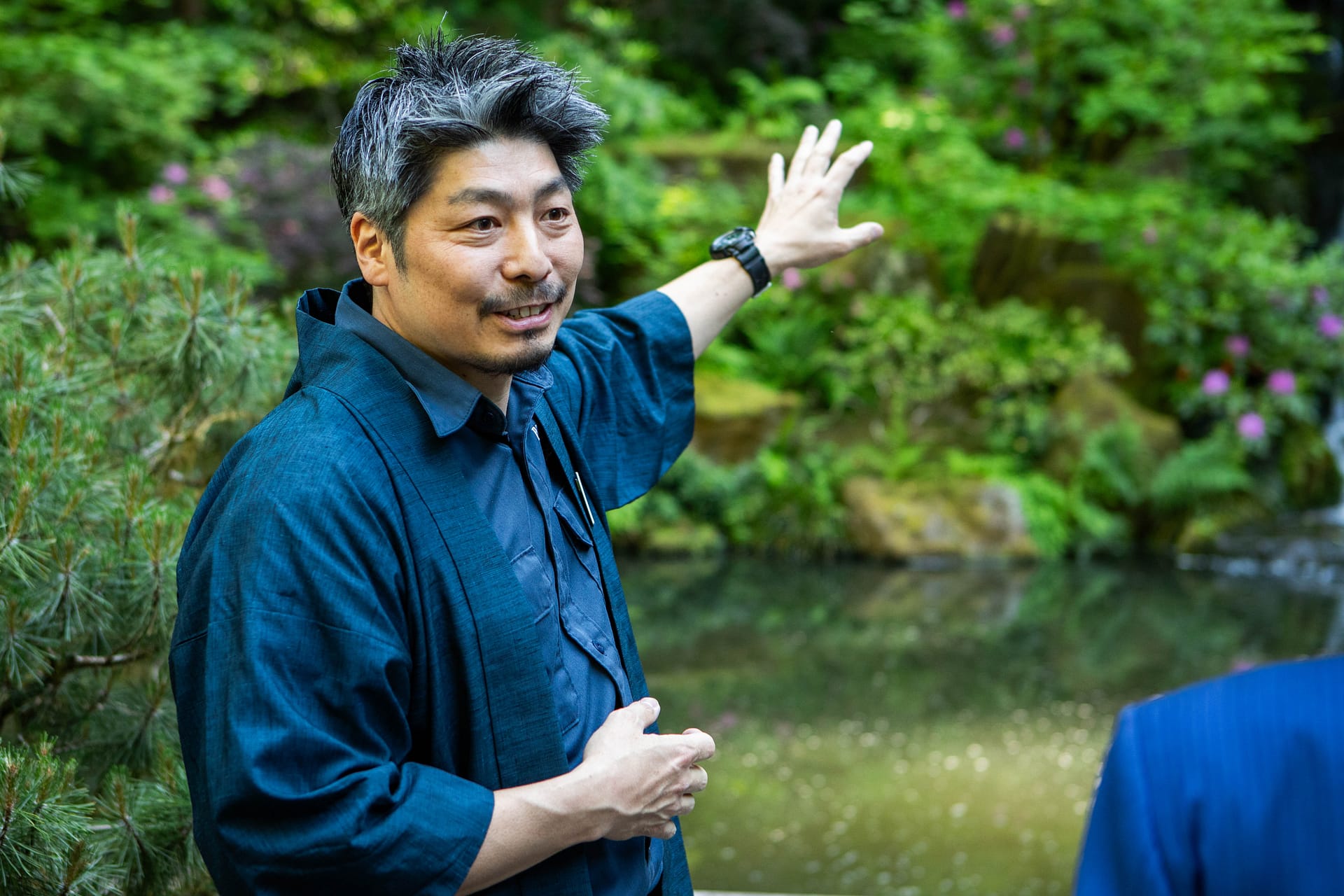 garden curator Hugo Torii stands smiling and gesturing to a waterfall wearing a Japanese gardening outfit and happi coat